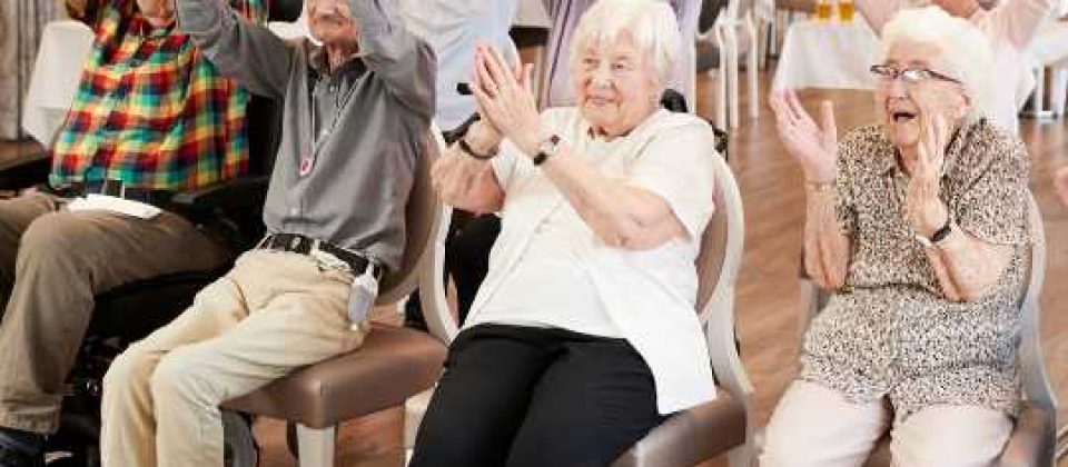 A group of older adults seated in chairs at a senior center, raising their arms and clapping together, participating in an interactive activity.