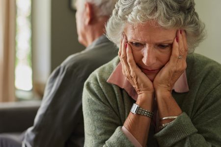 Older woman sitting with her head in her hands, looking upset, while an older man faces away in the background.