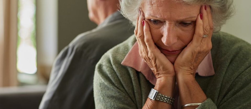 Older woman sitting with her head in her hands, looking upset, while an older man faces away in the background.