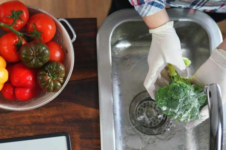 June 25′ Caregiver Newsletter Square Images 1 Person wearing gloves washing broccoli at a kitchen sink next to a bowl of fresh tomatoes and peppers.