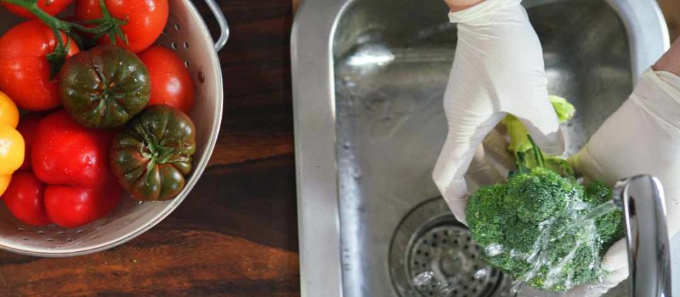 Person wearing gloves washing broccoli at a kitchen sink next to a bowl of fresh tomatoes and peppers.