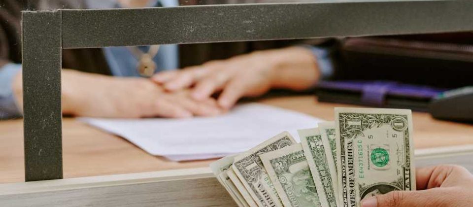 A person hands over a stack of dollar bills across a counter to an elderly woman who is partially visible behind a service window.