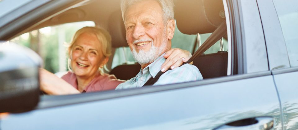 An older man in the drivers seat of a car wearing a seatbelt with an older woman sitting next to him.