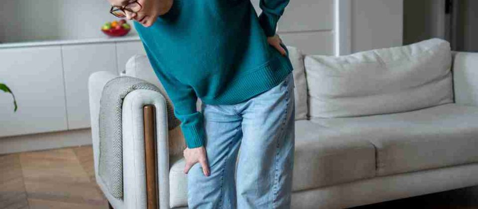 Person standing beside a couch and holding their lower back and leg, suggesting back or joint pain.