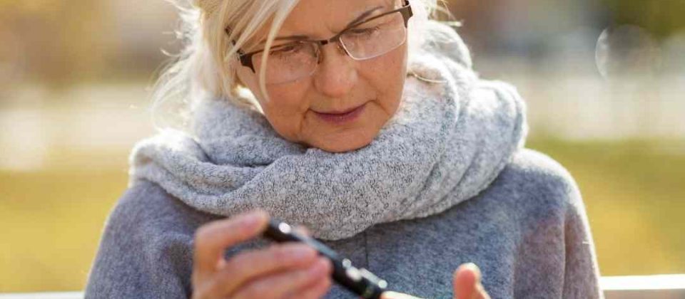 An elderly woman with white hair is sitting outside poking her finger with a lancing device to check her blood sugar.