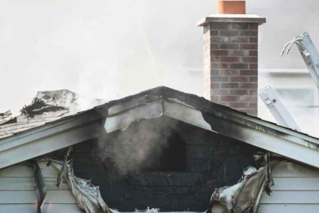Smoke rises from a fire-damaged house with burned siding and roof damage.