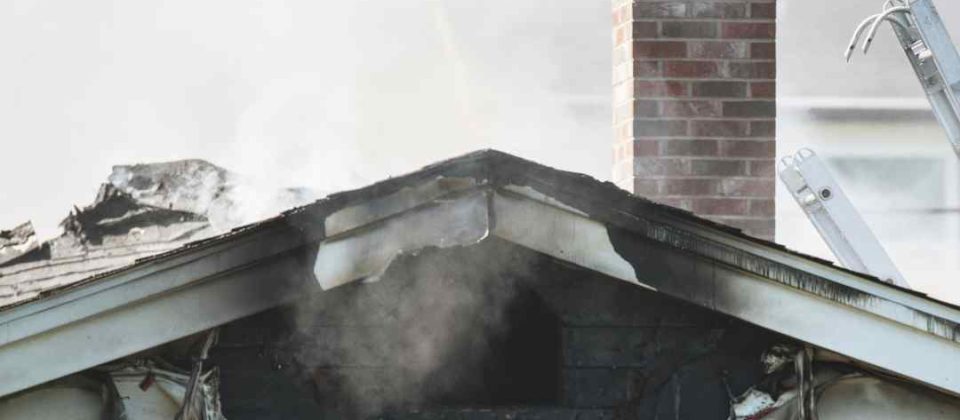 Smoke rises from a fire-damaged house with burned siding and roof damage.