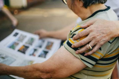 Older woman seated while looking at a photo album, with a hand resting on her shoulder.