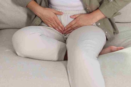 Person sitting on a couch with both hands placed over their lower abdomen, suggesting stomach pain or discomfort.