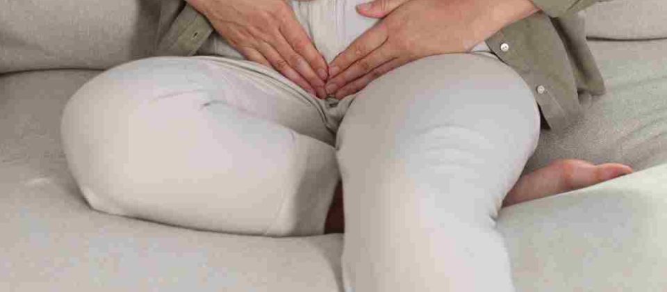 Person sitting on a couch with both hands placed over their lower abdomen, suggesting stomach pain or discomfort.