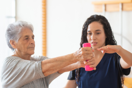 A physical therapist assists an elderly woman in arm exercises with a weight. The therapist supports her as she lifts the weight, promoting strength and mobility