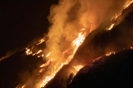 Wildfire burning across a hillside at night with heavy smoke and visible flames.