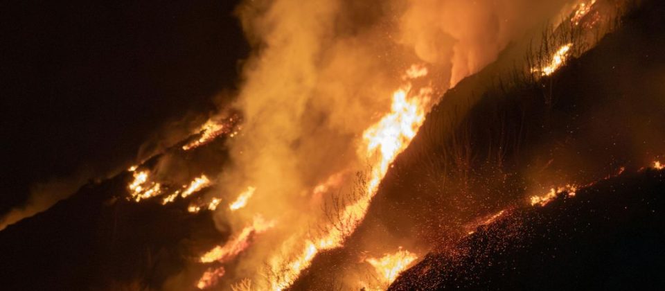 Wildfire burning across a hillside at night with heavy smoke and visible flames.