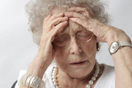 An elderly woman holds her head with a pained expression.