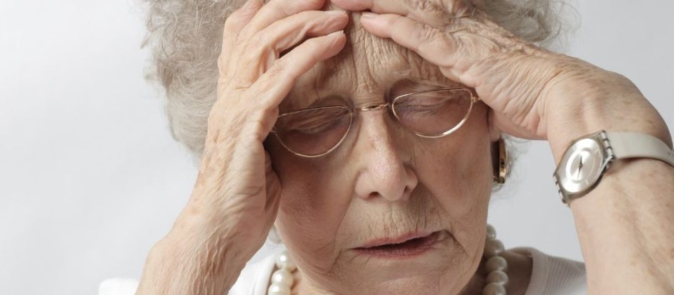 An elderly woman holds her head with a pained expression.