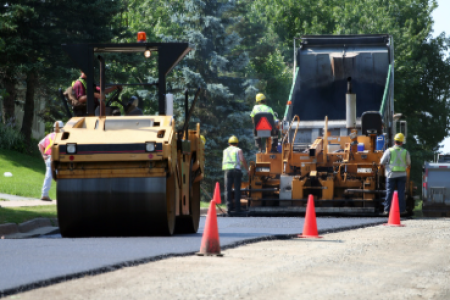 Road construction workers operate paving equipment while laying fresh asphalt on a roadway, with traffic cones marking the work area.