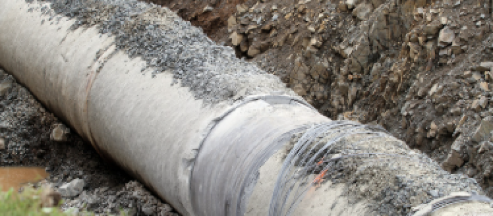 A large concrete pipe exposed in a dirt trench, with soil and gravel surrounding the pipe and grass visible in the foreground.