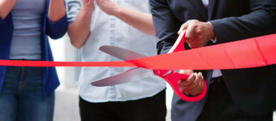Person in a suit cuts a red ceremonial ribbon with oversized scissors as attendees applaud.